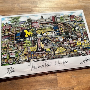 May include: A colorful illustration of a city scene with buildings, shops, and streets. The illustration is titled "Hail to the Pubs" of Ann Arbor. The illustration is signed by the artist, "Jay Kalena". The illustration is numbered 953/2500.