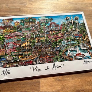 May include: A colorful illustration of a city scene with buildings, streets, and people. The title "Pubs of Miami" is written in black ink at the bottom of the print. The print is numbered 529/2500.