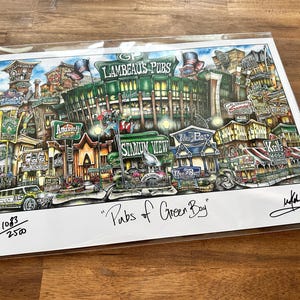 May include: A colorful print titled "Pubs of Green Bay" depicts a detailed street scene with buildings, signs, and a stadium. The artwork includes the text "Lambeau's Pubs" and is signed by the artist.