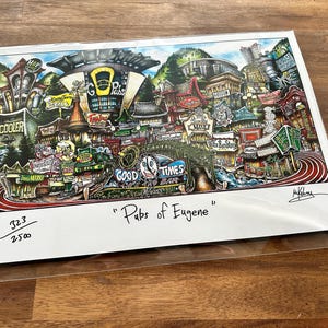 May include: A colorful print titled "Pubs of Eugene" featuring a detailed illustration of a city scene with various buildings, signs, and a mountain backdrop. The artwork is signed and numbered 323/2500.