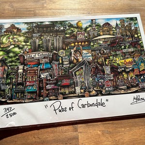 May include: A colorful illustration of a town with the title "Pubs of Carbondale" printed below. The illustration is a whimsical depiction of a town with buildings, streets, and people. The illustration is signed by the artist.
