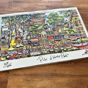 May include: A colorful art print titled Pubs of Winter Park featuring a detailed illustration of a bustling street scene with various storefronts, trees, and a clock tower. The print is numbered 254/2500 and includes a signature in the bottom right corner.