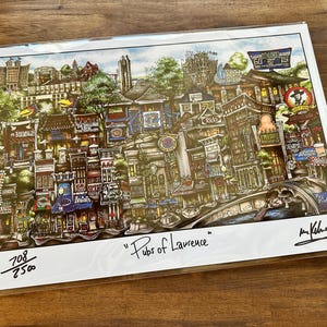 May include: A detailed color print titled Pubs of Lawrence featuring a dense cityscape of various buildings and storefronts with signage. The artwork includes a numbered edition of 708/2500 and a signature in the bottom right corner.