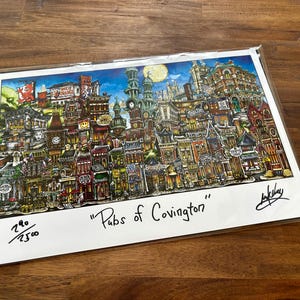 May include: A colorful print titled "Pubs of Covington" depicts a detailed illustration of a city street with numerous buildings, including pubs, under a blue sky with a full moon. The artwork is signed and numbered.