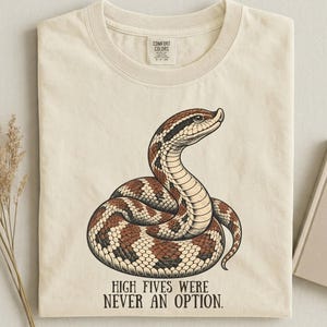 Può includere: Una t-shirt color crema con un'illustrazione grafica di un serpente marrone e nero avvolto su se stesso. Sotto il serpente, il testo recita: "HIGH FIVES WERE NEVER AN OPTION." La maglietta ha un girocollo e un'etichetta del marchio Comfort Colors.