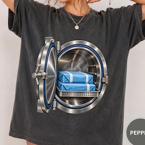 May include: A charcoal gray short-sleeve t-shirt featuring a large graphic of an open circular metal vault door. Inside the vault, two blue rectangular packages are stacked on a metal shelf. The word PEPPER appears in a small gray circle.