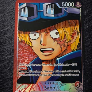 May include: A One Piece trading card featuring the character Sabo with blonde hair and blue goggles. The card has a red and black background with text detailing game rules, a power level of 5000, and the label Leader Sabo Dressrosa/Revolutionary Army.