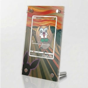 Könnte beinhalten: Ein transparenter Acryl-Displayrahmen mit einer japanischen Rowlet-Pokémon-Sammelkarte. Der Rahmen zeigt einen Hintergrund, der von Der Schrei inspiriert ist, mit orangefarbenen, gelben und dunkelgrünen wirbelnden Linien. Vier silberne Metallschrauben fixieren die Karte.