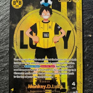 May include: A rectangular trading card featuring an anime character wearing a yellow and black soccer jersey with the BVB 09 logo, balancing a soccer ball on their head. The background has yellow and black vertical stripes with the text Monkey D. Luffy.