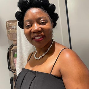 May include: A woman wearing a black dress with a beaded necklace and earrings. Her hair is styled in a unique updo. The dress has a sweetheart neckline and thin straps.