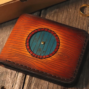 Op de afbeelding: Een bi-fold portemonnee met een bruin houtnerfpatroon en een centraal cirkelvormig blauwgroen paneel. De blauwgroene cirkel is omgeven door een rode rand en heeft een kleine messingkleurige knop in het midden.