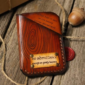 May include: A handcrafted brown leather wallet with a wood grain design and the quote "What has it got in its nasty little pocketses?" It also features a "no admittance except on party business" tag. A gold ring and acorn are nearby.