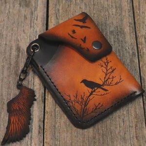 Puede incluir: Una cartera de cuero con acabado degradado en naranja y negro, con un gráfico de un pájaro negro posado en una rama. La cartera incluye un cierre a presión, una anilla metálica negra y un colgante de cuero extraíble en forma de ala.