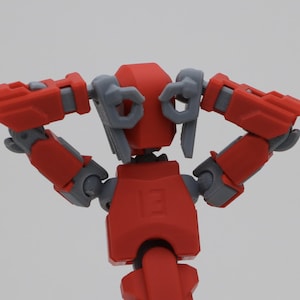 May include: A red and grey articulated robot action figure with the number 13 embossed on its chest. The figure is posed with its arms bent at the elbows and hands positioned behind its head, showcasing mechanical joints and geometric armour plates.
