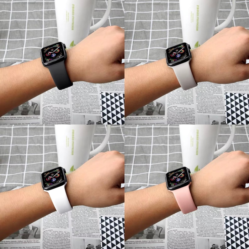 May include: Four Apple Watches with different colored bands: black, off-white, white, and pink. Each watch displays a colorful screen and is worn on a wrist. The bands are made of a smooth material. The background includes a newspaper and a white mug.