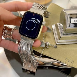 May include: A silver-toned smartwatch with a unique, flowing metal band. The watch face displays the time, 19:16, in white digits against a dark background. A silver chain with a spherical charm is also visible.