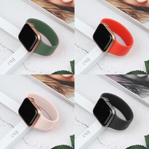 May include: Four smartwatches with different colored bands: green, red, pink, and black. Each watch has a black screen and a metallic case. The bands are made of a smooth material and are displayed on a white surface.