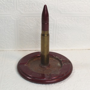 May include: A unique ashtray featuring a repurposed artillery shell. The shell is a brass color with a maroon tip, set in a circular, maroon-colored base. The base has a recessed area for holding ashes.