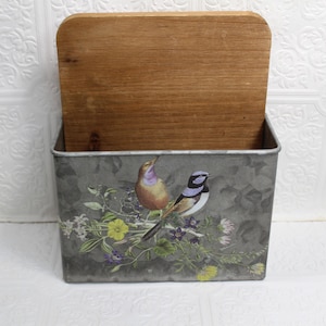 May include: A galvanized metal wall-mounted storage bin with a hinged wooden lid. The front of the bin features a colorful illustration of two birds perched on flowering branches. The bin is designed for organizing and storage.