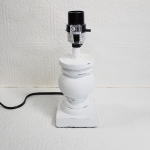 May include: A white table lamp with a distressed finish. The lamp has a black lamp holder and a black power cord. The base is square, and the body has a rounded shape. The lamp is designed for indoor use.