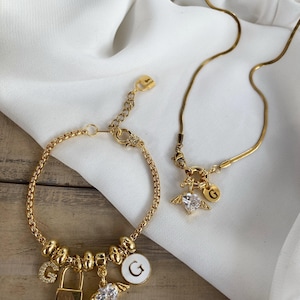 Pode incluir: Um conjunto de colar e pulseira de corrente dourada com um pingente de coração com asas e um pingente de letra G. A pulseira tem um pingente de cadeado e um pingente de letra G.