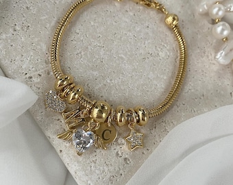 May include: A gold charm bracelet featuring a selection of charms, including a winged heart, a star, a letter 'C', and a heart with the letter 'M'.