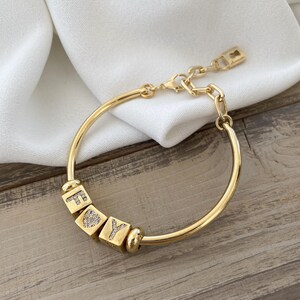 Puede incluir: Pulsera dorada con las letras "FOY" en cuentas cuadradas. La "O" está representada por un corazón. La pulsera tiene un cierre de cadena y broche. El fondo es una tela blanca y una superficie de madera.