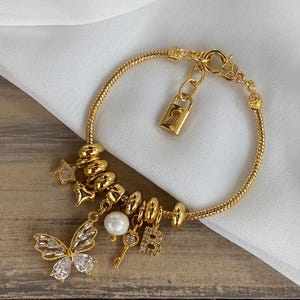 Op de afbeelding: Een gouden armband met bedels, waaronder een vlinder, een sleutel, een parel, een ster, een hart en een letter "B". De armband heeft ook een gouden slot en sluiting.