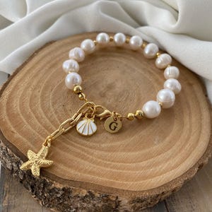 May include: A gold bracelet with white pearls, a gold starfish charm, a gold seashell charm, and a gold charm with the letter "G".