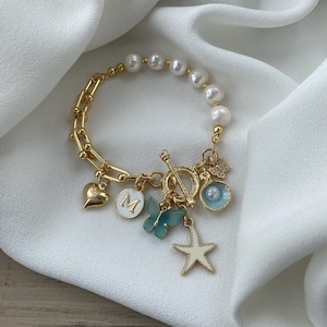 Op de afbeelding: Een gouden armband met witte parels, een hartvormige bedel, een vlinderbedel, een stervormige bedel, een schelpbedel en een bedel met de letter "M".