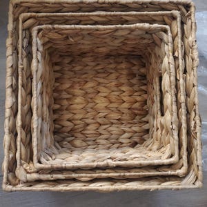May include: A set of three woven rectangular baskets in varying sizes. The baskets are made of a light brown, natural material, with a textured surface. The baskets are nested inside each other, showcasing their graduated sizes and open tops.