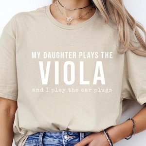 May include: A beige t-shirt with the text "My daughter plays the viola and I play the ear plugs".