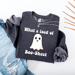 Puede incluir: Una sudadera gris oscuro con la frase "What a load of Boo-Sheet" y un dibujo de un fantasma. Un collar plateado está colocado sobre la sudadera, que está doblada sobre unos vaqueros azules.