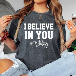 Könnte beinhalten: Ein dunkelgraues T-Shirt mit dem Text "I BELIEVE IN YOU #testday" in Weiß.