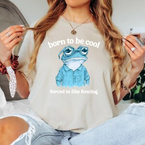 May include: A light beige t-shirt with a blue frog wearing glasses and a blue shirt. The text "born to be cool" and "forced to like fencing" is printed on the shirt.