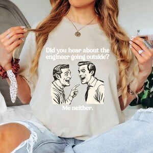 May include: A light beige t-shirt featuring a vintage-style illustration of two men talking. The text on the shirt reads "Did you hear about the engineer going outside? Me neither."