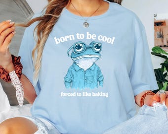 Op de afbeelding: Lichtblauw T-shirt met een cartoon kikker die een zonnebril draagt en de tekst "born to be cool forced to like baking".