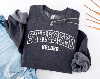 May include: A charcoal grey sweatshirt featuring the text "STRESSED WELDER" in white, block lettering. The sweatshirt is folded, alongside light blue ripped jeans and a silver necklace. The Gildan brand label is visible.
