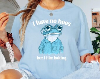 Op de afbeelding: Lichtblauw T-shirt met een cartoon kikker met een bril en de tekst "I have no hoes but I like baking".