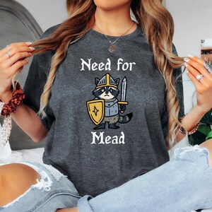 Op de afbeelding: Donkergrijs t-shirt met de tekst "Need for Mead" boven een cartoon wasbeer met een helm en een zwaard en schild. De wasbeer draagt een ridderoutfit.