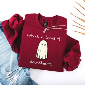 Puede incluir: Una sudadera granate con la frase "What a load of Boo-Sheet" y un dibujo de un fantasma. La sudadera se combina con unos vaqueros azules y un collar plateado. El diseño está impreso en blanco.