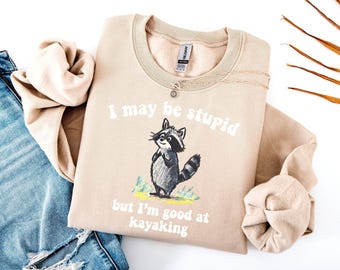 Puede incluir: Sudadera beige con el texto "I may be stupid but I'm good at kayaking" y un dibujo de mapache. La sudadera está doblada y colocada junto a unos vaqueros azules.