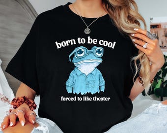 May include: Black t-shirt with a blue frog wearing glasses and a blue shirt. The text "born to be cool" and "forced to like theater" is printed on the shirt.