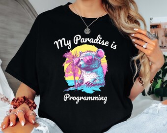 Op de afbeelding: Zwart T-shirt met een afbeelding van een wasbeer met een zonnebril en een palmboom. De tekst "My Paradise is Programming" staat op het T-shirt gedrukt.