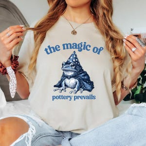 May include: A beige t-shirt with a blue frog wearing a blue wizard hat and a blue cape. The text "the magic of pottery prevails" is printed on the shirt.
