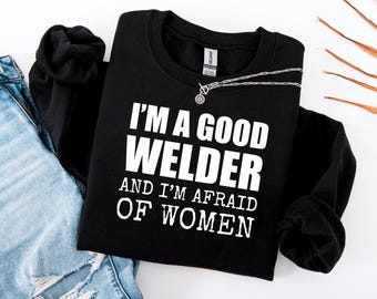 May include: Black crew neck sweatshirt with the white text "I'M A GOOD WELDER AND I'M AFRAID OF WOMEN". A silver necklace with a circular pendant is on the sweatshirt. The sweatshirt is next to blue jeans.