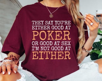 May include: A maroon t-shirt with the text "THEY SAY YOU'RE EITHER GOOD AT POKER OR GOOD AT SEX I'M NOT GOOD AT EITHER" in white and orange lettering. The shirt is a casual style.