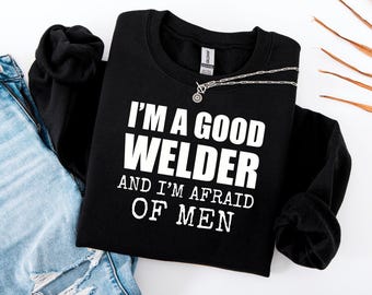 May include: Black crew neck sweatshirt with the white text "I'M A GOOD WELDER AND I'M AFRAID OF MEN". A silver necklace with a circular pendant is on the sweatshirt. The sweatshirt is next to blue denim jeans.