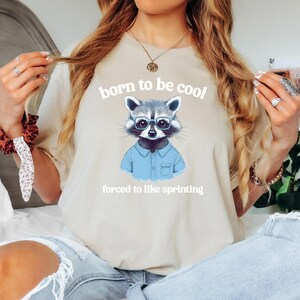 Puede incluir: Una camiseta beige claro con un gráfico de un mapache con gafas azules y una camisa azul de botones. El texto "born to be cool" está por encima del mapache y el texto "forced to like sprinting" está por debajo del mapache.