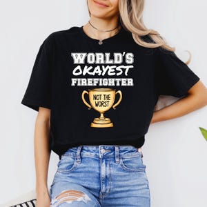 May include: Black t-shirt with the text "WORLD'S OKAYEST FIREFIGHTER" in white block letters. Below the text is a gold trophy with the words "NOT THE WORST". The shirt is worn with blue jeans.
